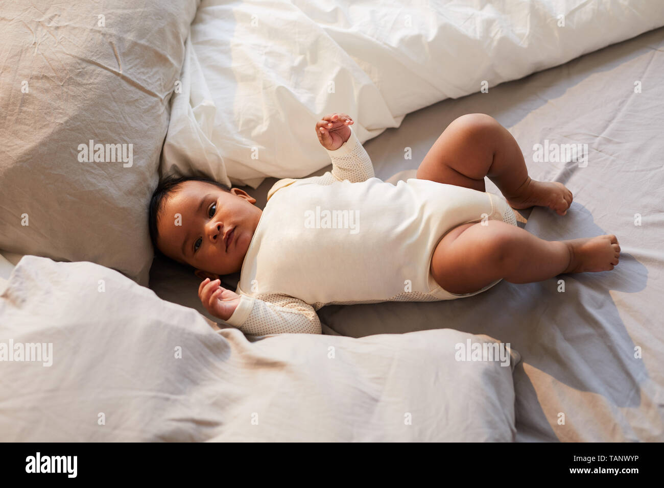 Portrait of serious adorable African baby boy in bodysuit lying between pillows in comfortable bed Stock Photo