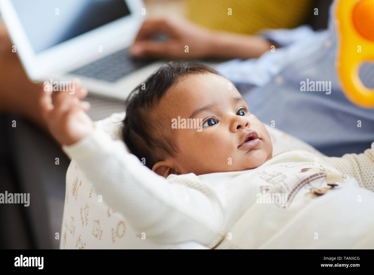 Close-up of content curious baby boy in bodysuit lying in cocoon and looking at rattle while mother working with laptop in background Stock Photo