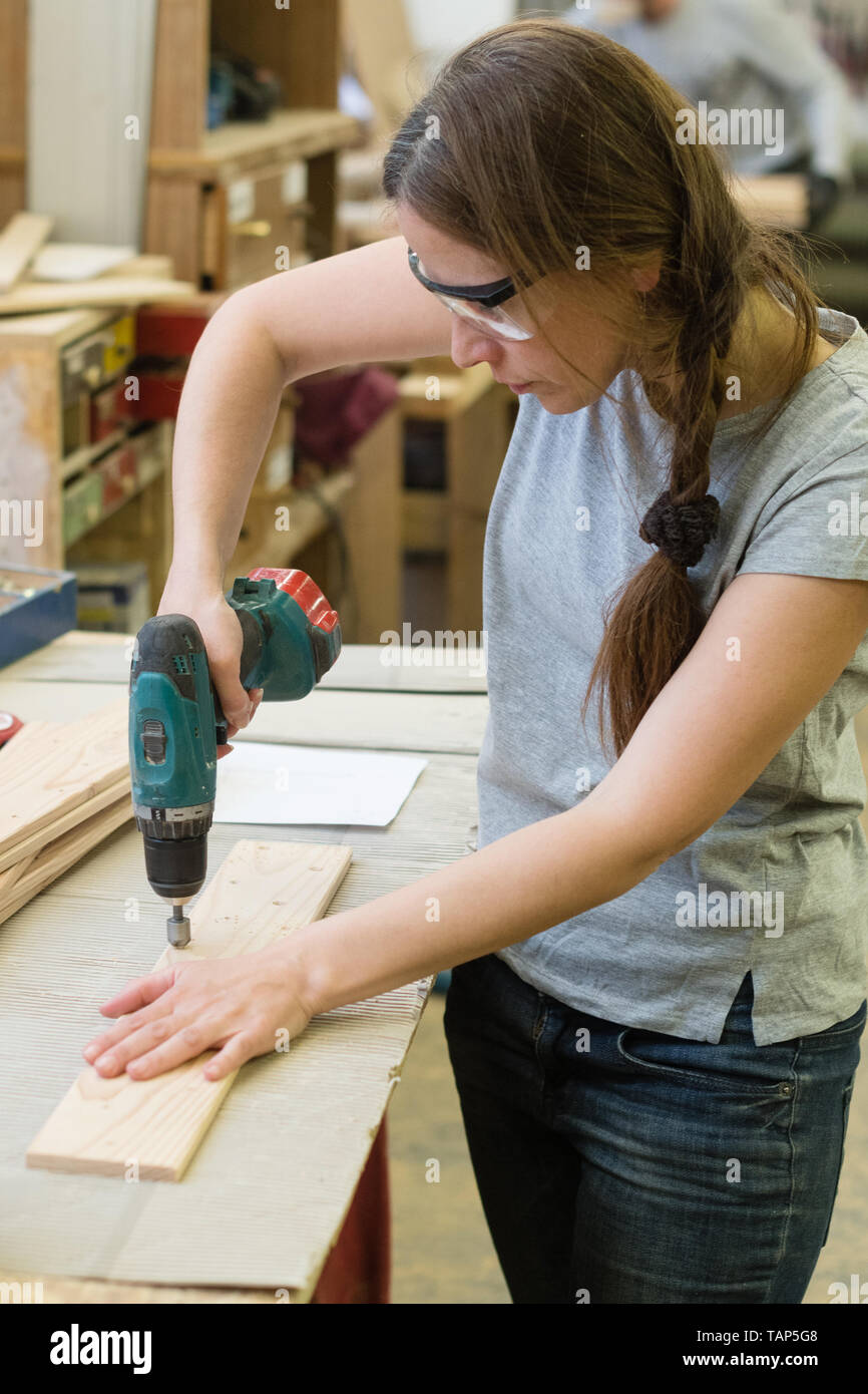 Young woman with drill making hole into wooden plank at workshop Stock Photo