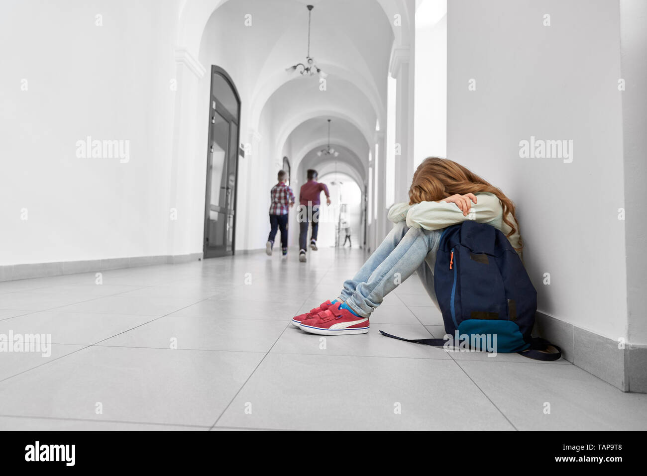 Girl with long hair in shirt, jeans and sneakers leaning on knees. Lonely, sad schoolgirl sitting on grey floor with bending head. Shool backpack near child. Stock Photo