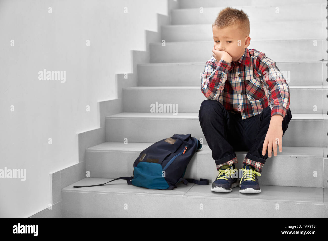 Handsome, thoughtful schoolboy sitting on stairwell in school, leaning head on hand. Lonely, sad, depressed child in checked shirt, jeans and sneakers thinking. Stock Photo