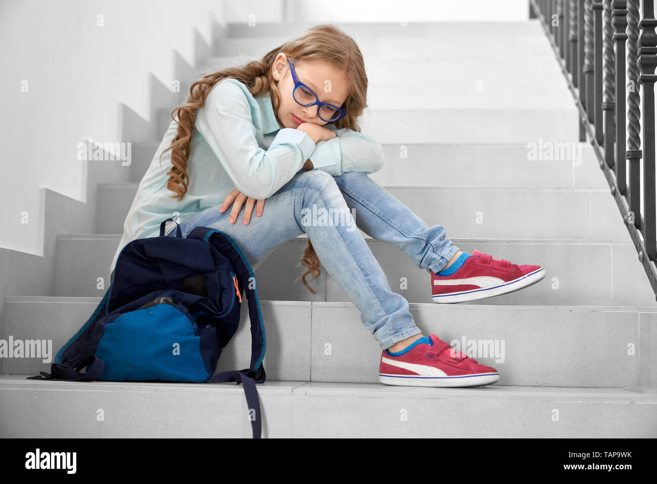 Pretty, sad schoolgirl in glasses sitting on staircase in school, leaning on knees. Depressed girl with long curly hair looking down, thinking. Stock Photo