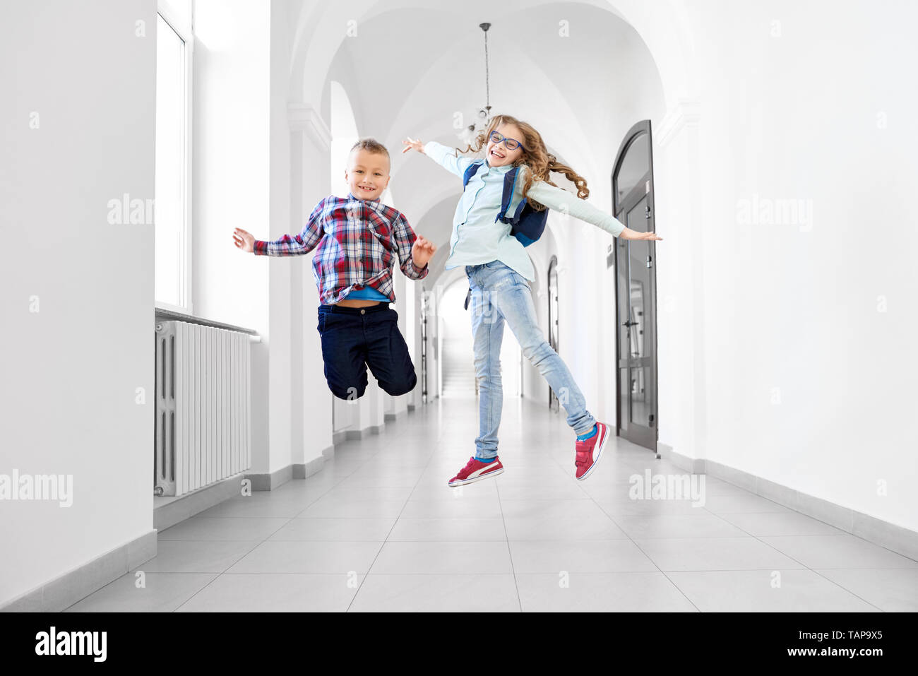 Happy, cheerful schoolchildren having fun after lessons in school hallway. Pretty, positive girl with school backpack and handsome boy smiling and jumping. Stock Photo