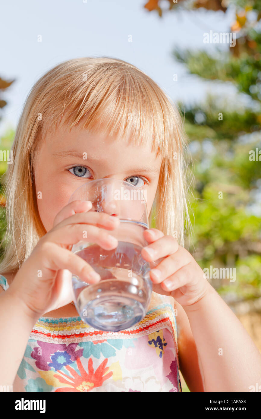 Blonde little girl drinking fresh and pure tap water from a glass in a summer garden - healthy lifestyle concept Stock Photo