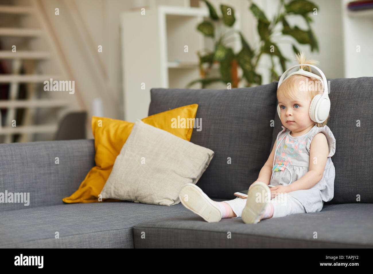 Serious curious little girl digital nomad in wired headphones sitting on comfortable sofa and listening to music via smartphone app Stock Photo