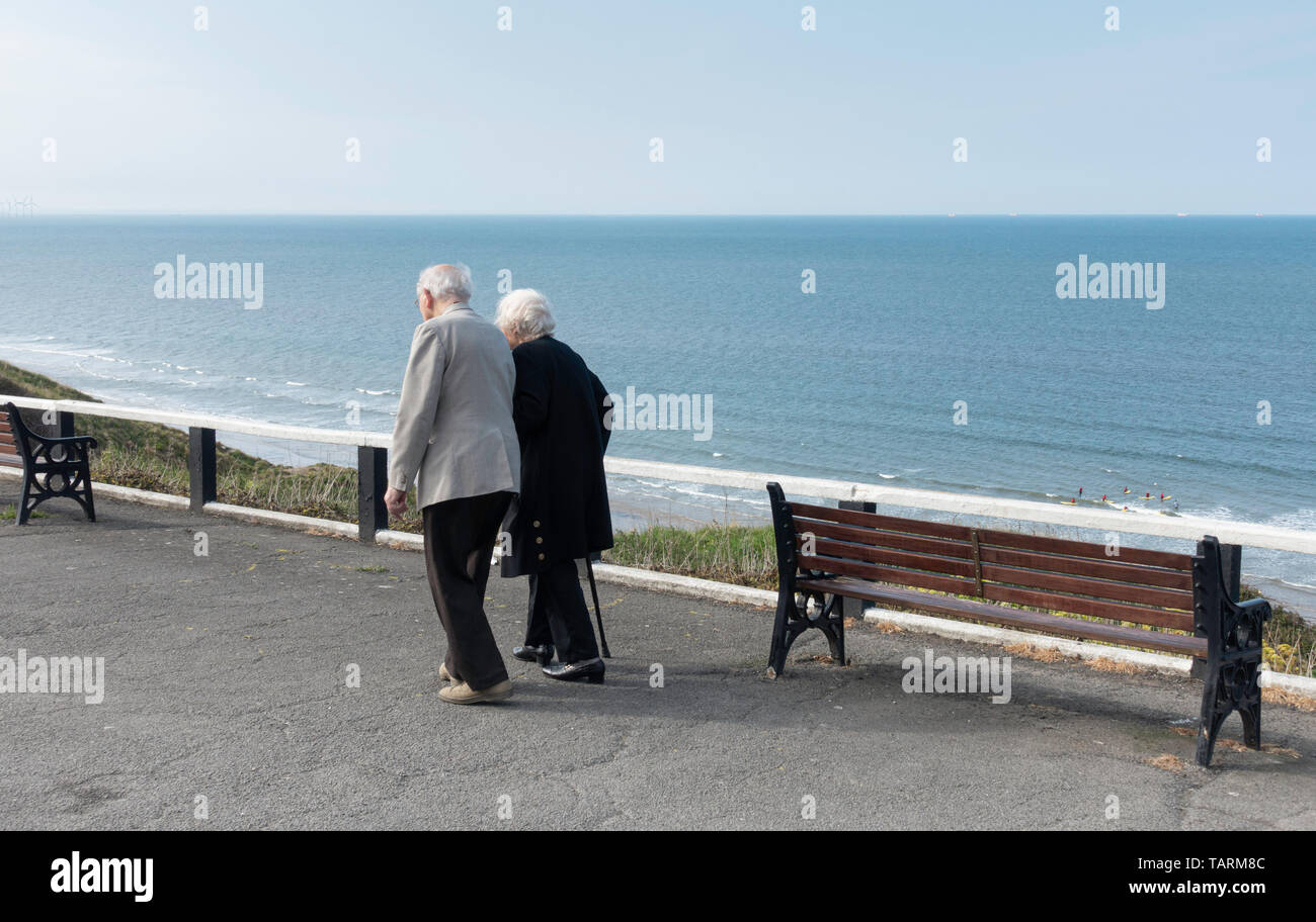 Elderly couple walking along promenade overloking the sea. UK Stock Photo
