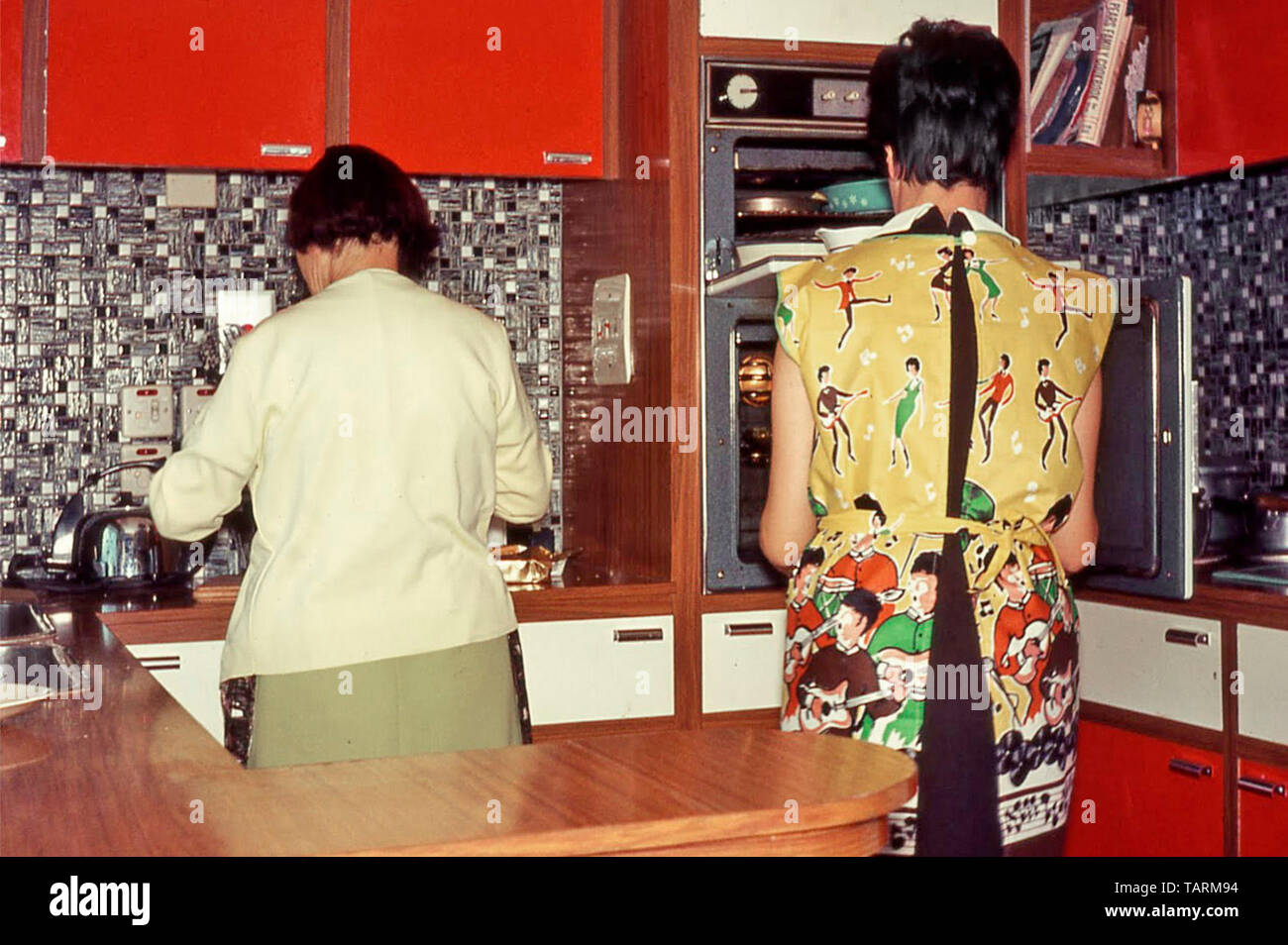 Archival back view of young model released housewife at oven & her  Mum prepare food in new 1966 DIY property released kitchen interior Formica worktops mother and daughter England UK Stock Photo