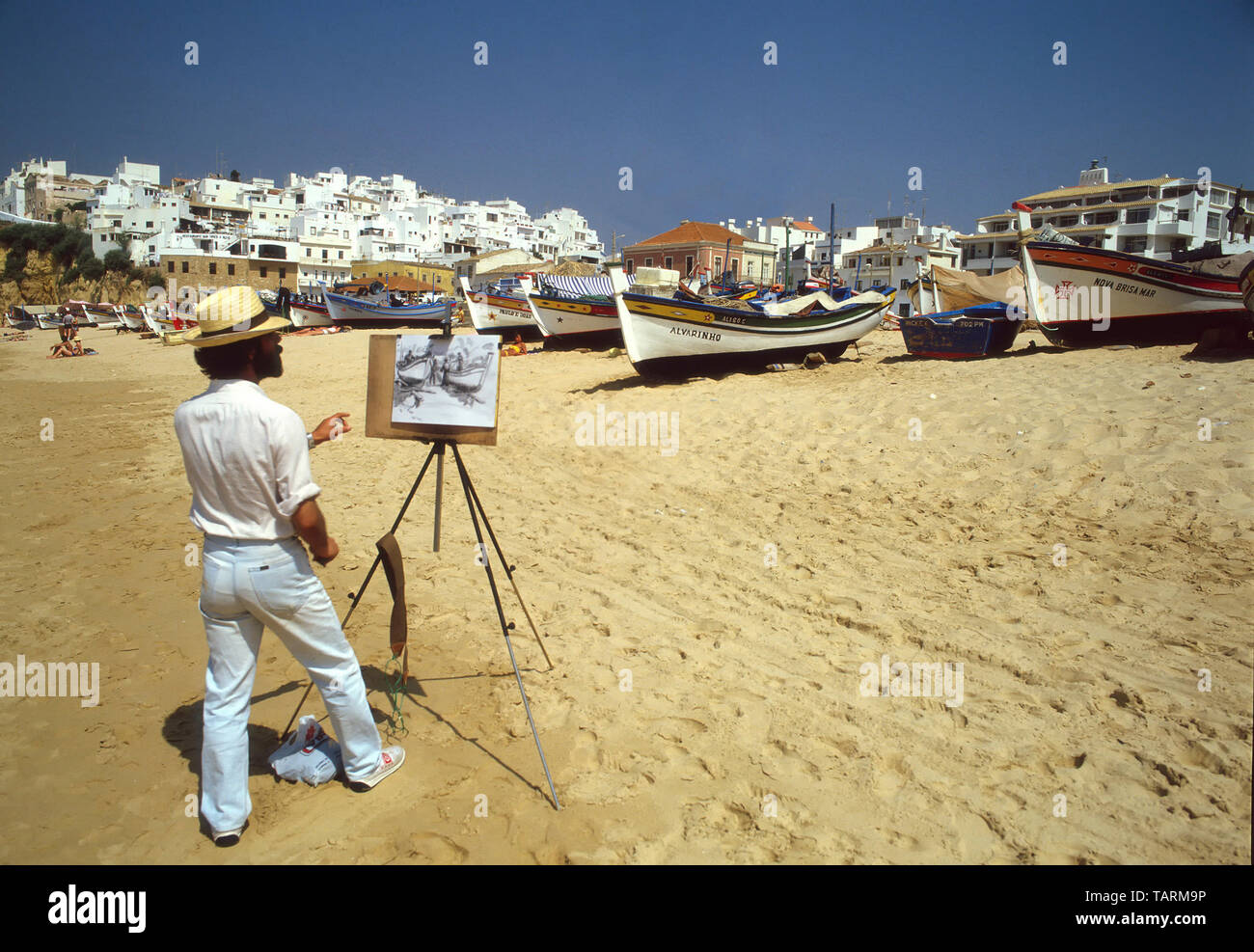 Man1980s artist at work under clear blue sky with easel sketching on fishing boat & holiday sunbathing clean sand beach Albufeira Algarve Portugal Stock Photo