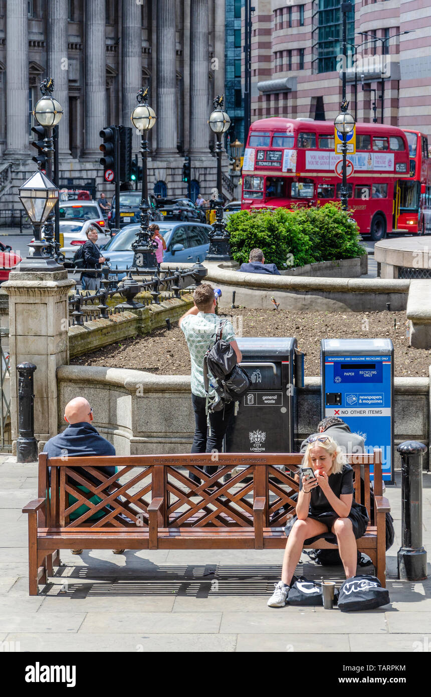 Tourists sit on wooden benches at Royal Exchange in London. A lady stops and looks at her mobile phone. Stock Photo