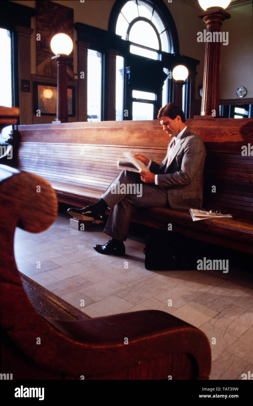 Businessman in gray suit reads financial newspaper while waiting for train in railway terminal waiting room. Stock Photo