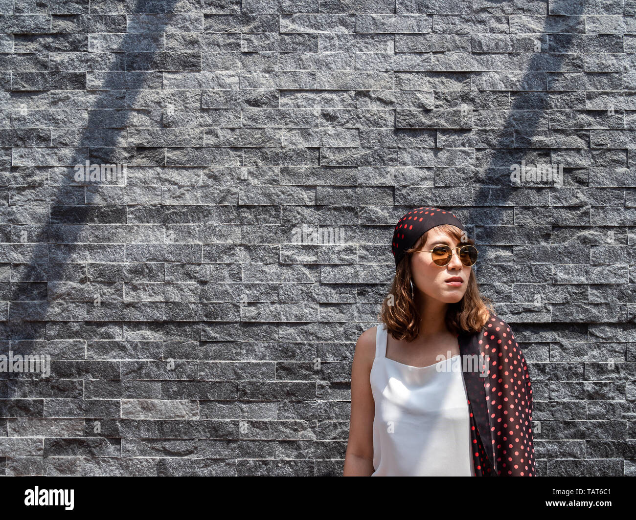 Beautiful fashionable hipster asian woman wearing white sleeveless, sunglasses and red polka dot pattern turban smiling on modern grey brick wall back Stock Photo