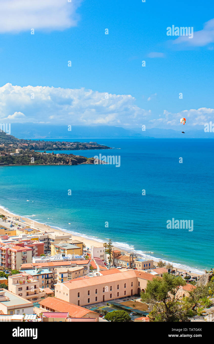 Paraglider flying over the beautiful blue bay in Italian Cefalu, Sicily captured on a vertical picture. Paragliding is a popular adventure sport. Stock Photo