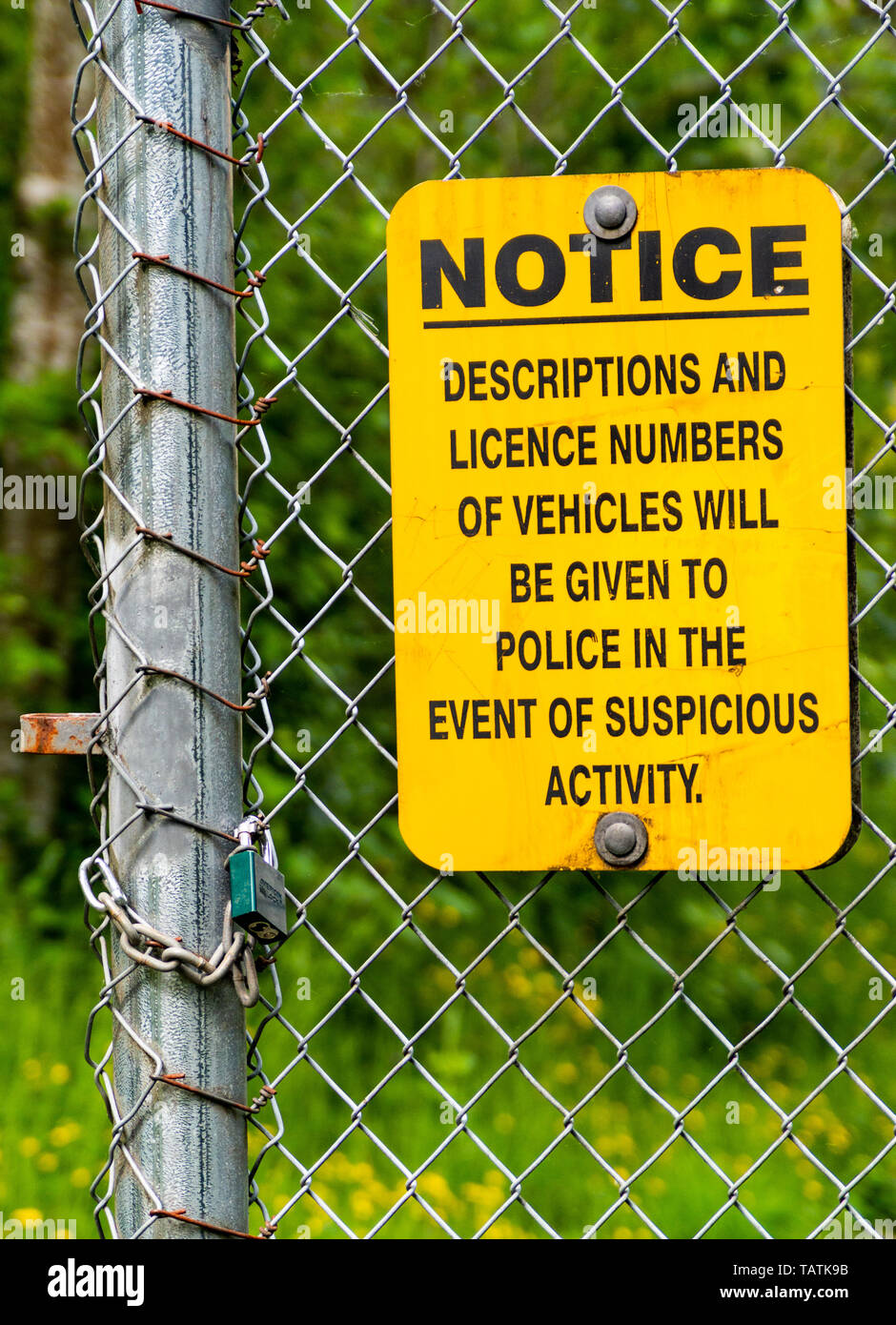 Yellow and black neighborhood crime and suspicious activity watch warning sign posted on chain link fence with wooded background beside open padlock.. Stock Photo