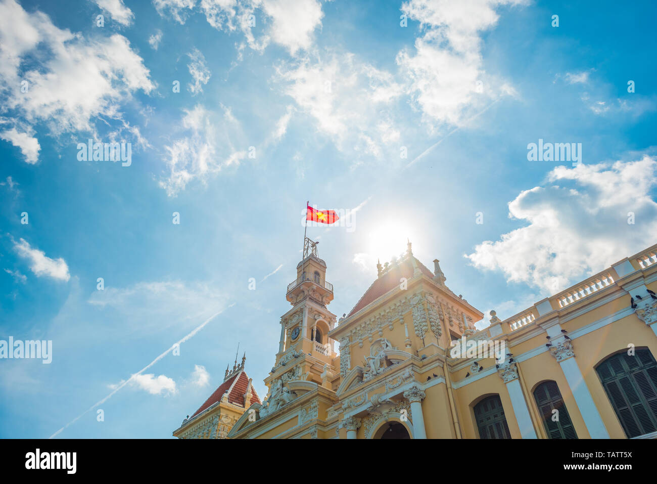 Saigon City Hall with waving Vietnamese flag against blue sky and curly summer clouds, sun, and airplane trail. Stock Photo
