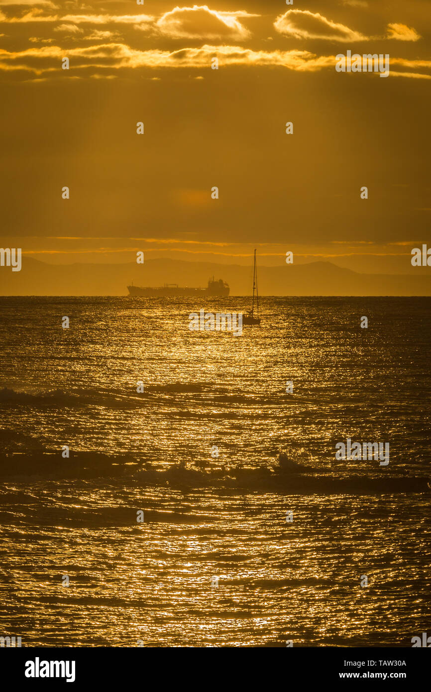 Portland, Dorset, UK. 28th May, 2019. A sailboat and tanker on the horizon as dawn breaks over the Dorset coast filling the sky with a golden light. Cloud and sun bring unsettled weather for today. Peter Lopeman/Alamy Live News Stock Photo