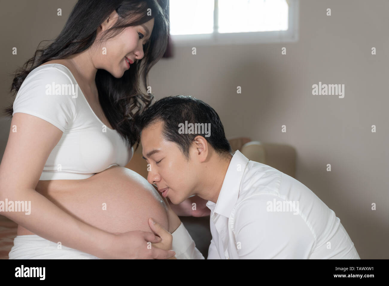 Asian couple pregnant wife and husband kissing on belly woman in bedroom background Stock Photo
