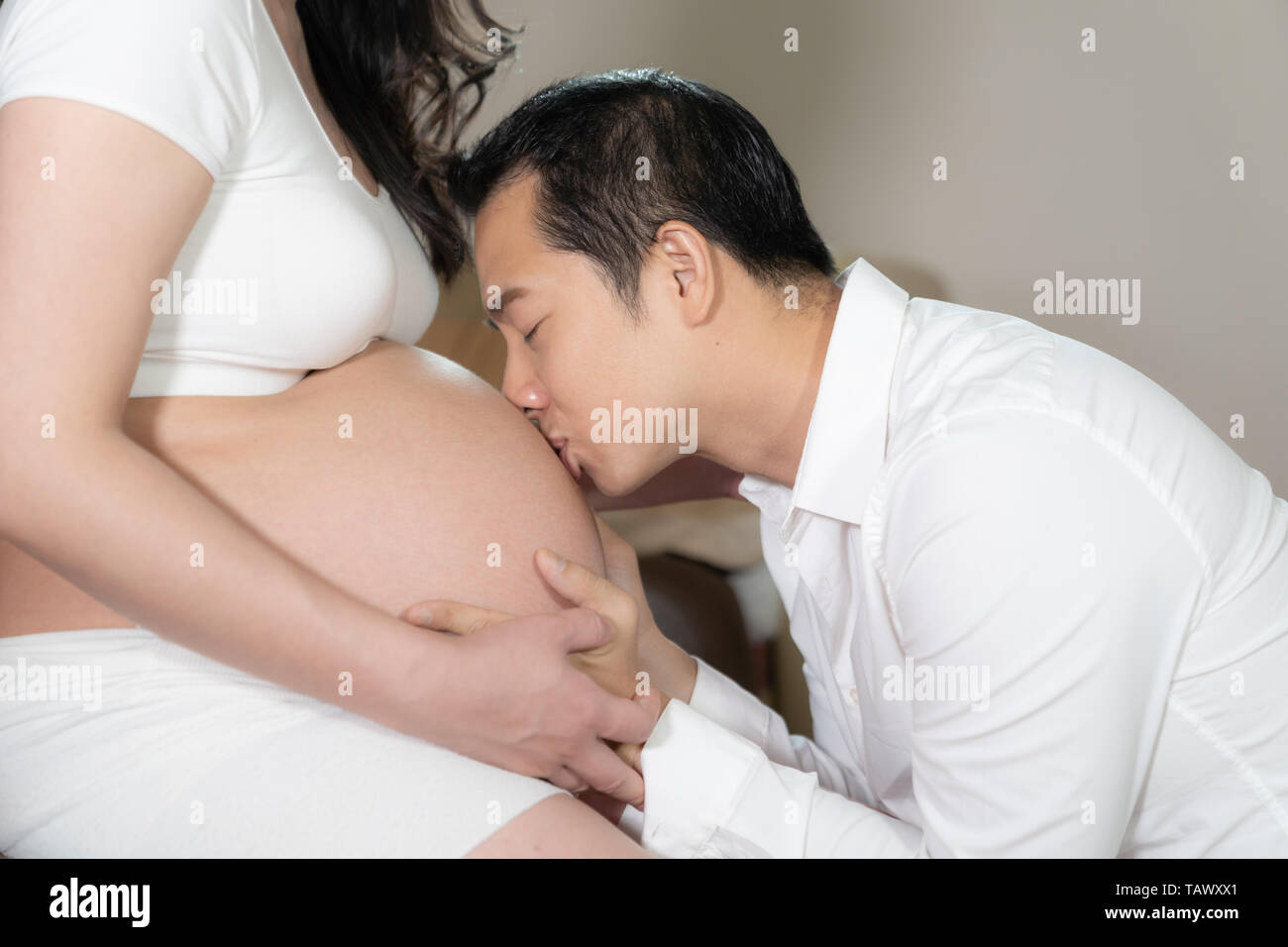 Asian couple pregnant wife and husband kissing on belly woman in bedroom background. Stock Photo