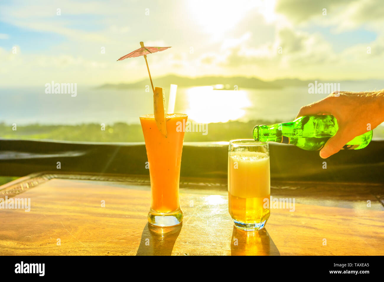 Male hand pours beer into a glass. Fruit cocktail and glass of beer on deck on foreground at sunset light. Spectacular view of Indian Ocean and Stock Photo
