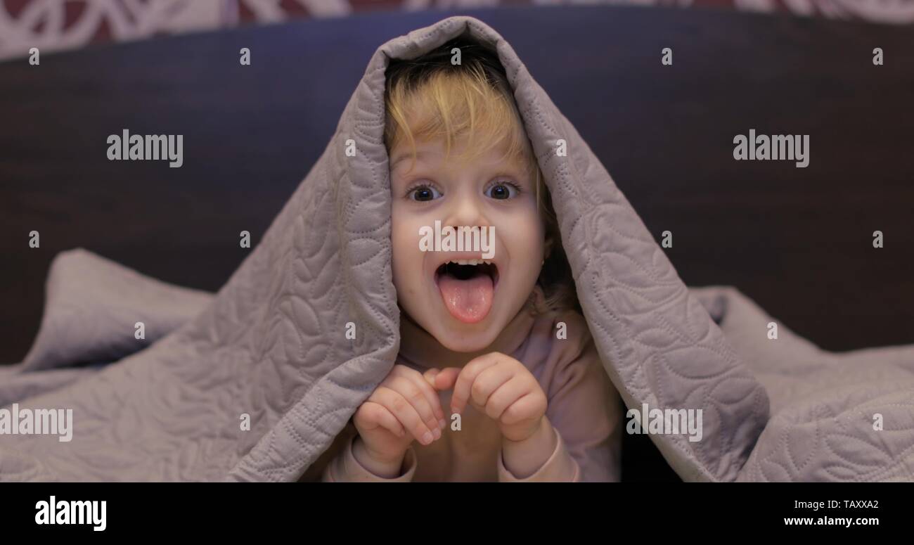 Little cheerful girl hides under a blanket and watching TV. Sweet