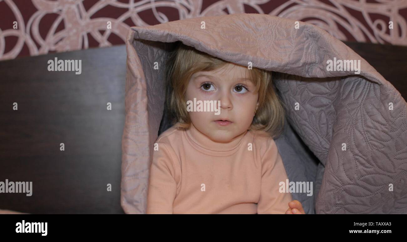 Little cheerful girl hides under a blanket and watching TV. Sweet, adorable child having fun on the bed under a coverlet. Concept of kids sleep, care or childhood Stock Photo