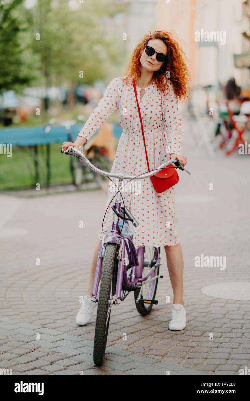 Vertical shot of curly red haired woman rides bicycle in city during weeked, has free time, wears long dress, shades and sneakers, carries small red b Stock Photo