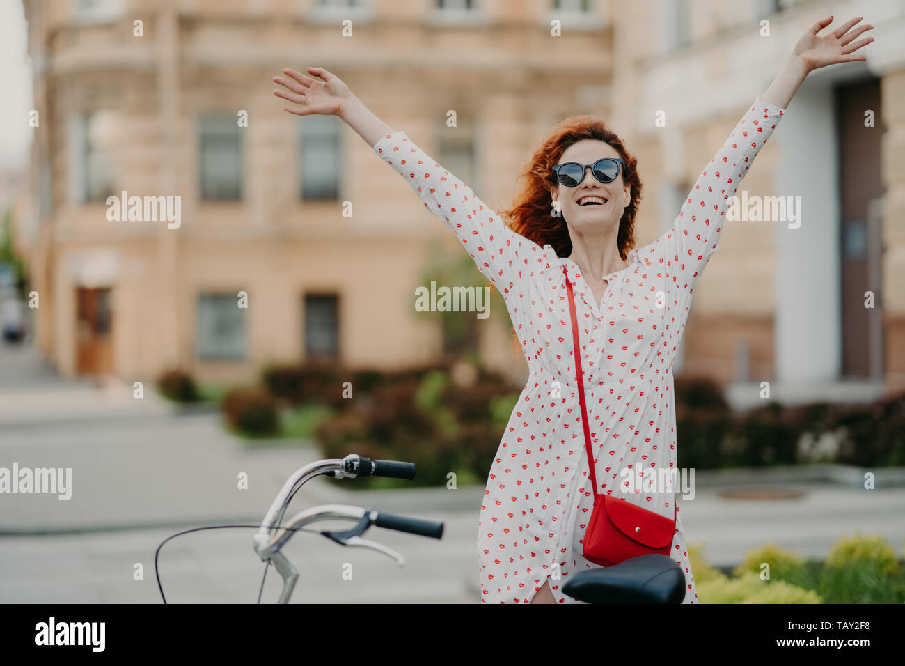 Horizontal outdoor shot of delighted positive red haired woman raises hands, feels free, dressed in summer clothes, rides bicycle on streets, enjoys r Stock Photo