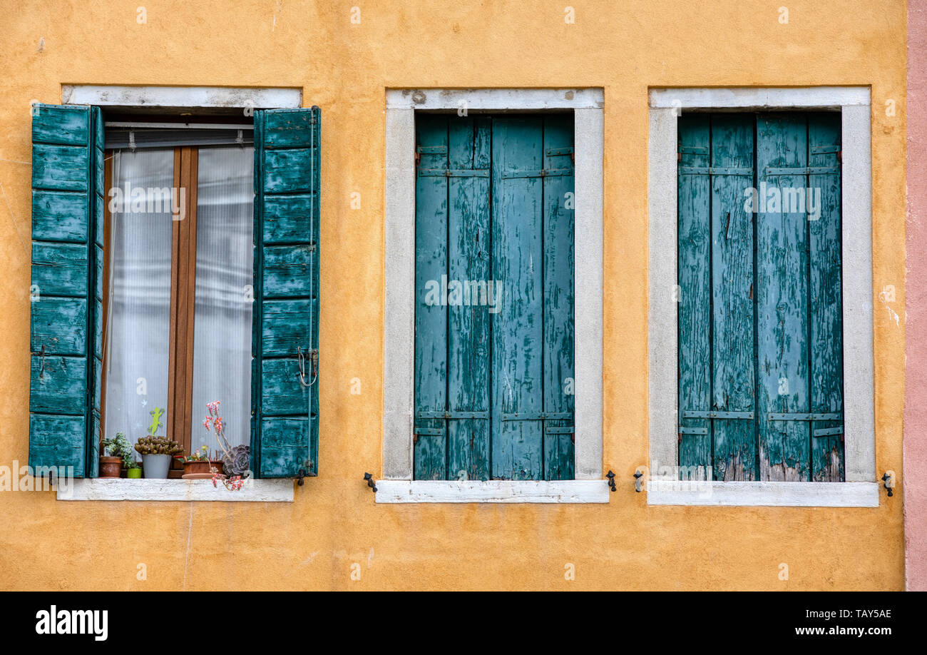 Photograph taken of three weathered, green painted windows against a textured yellow plaster wall in the medieval water town of Venice, Italy. Stock Photo