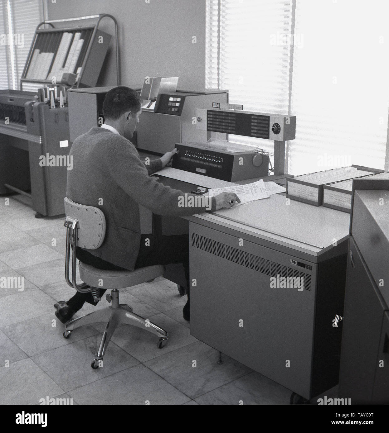 Mid 1960s, historical, a man working at the console of an IBM 1130 computing system, with other peripheral equipment in the room. Introduced in 1965, the 1130 was aimed at education and engineering and scientific markets. The 1131 central processing unit (CPU) was the primary processing component of the IBM 1130 and was built into a free-standing, desk-like enclosure, seen on the right of the operator in the picture. The CPU was based on hard-coded logic (SLT) modules and 'core memory', the highest-performing memory device available at the time. Stock Photo