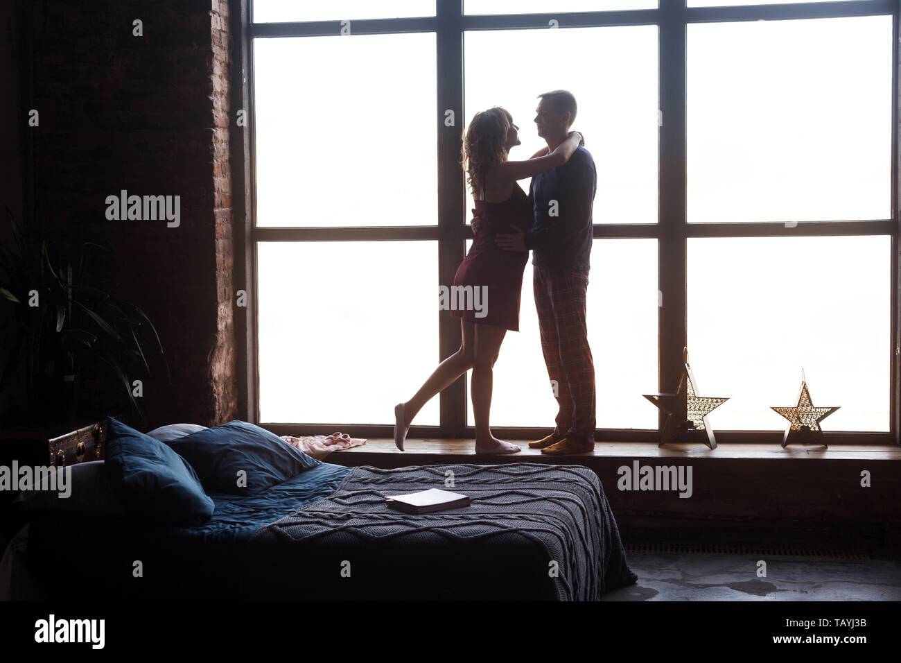 Silhouette of young happy beautiful couple expecting baby standing together in the big window sill hugging and looking to each other with love. Studio Stock Photo