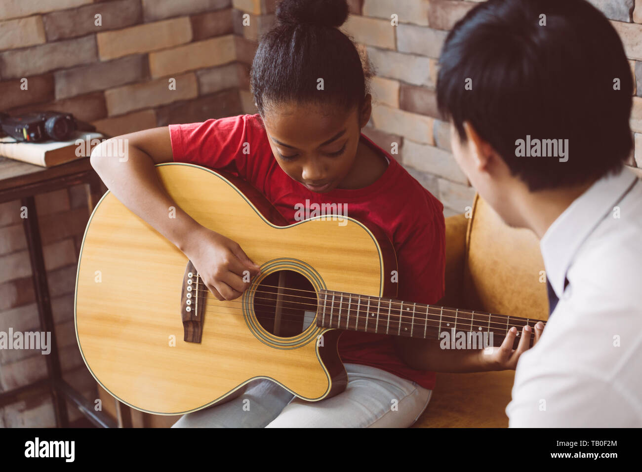 African American girl in casual outfit learning how to play guitar while sitting on couch near male tutor during music lesson Stock Photo