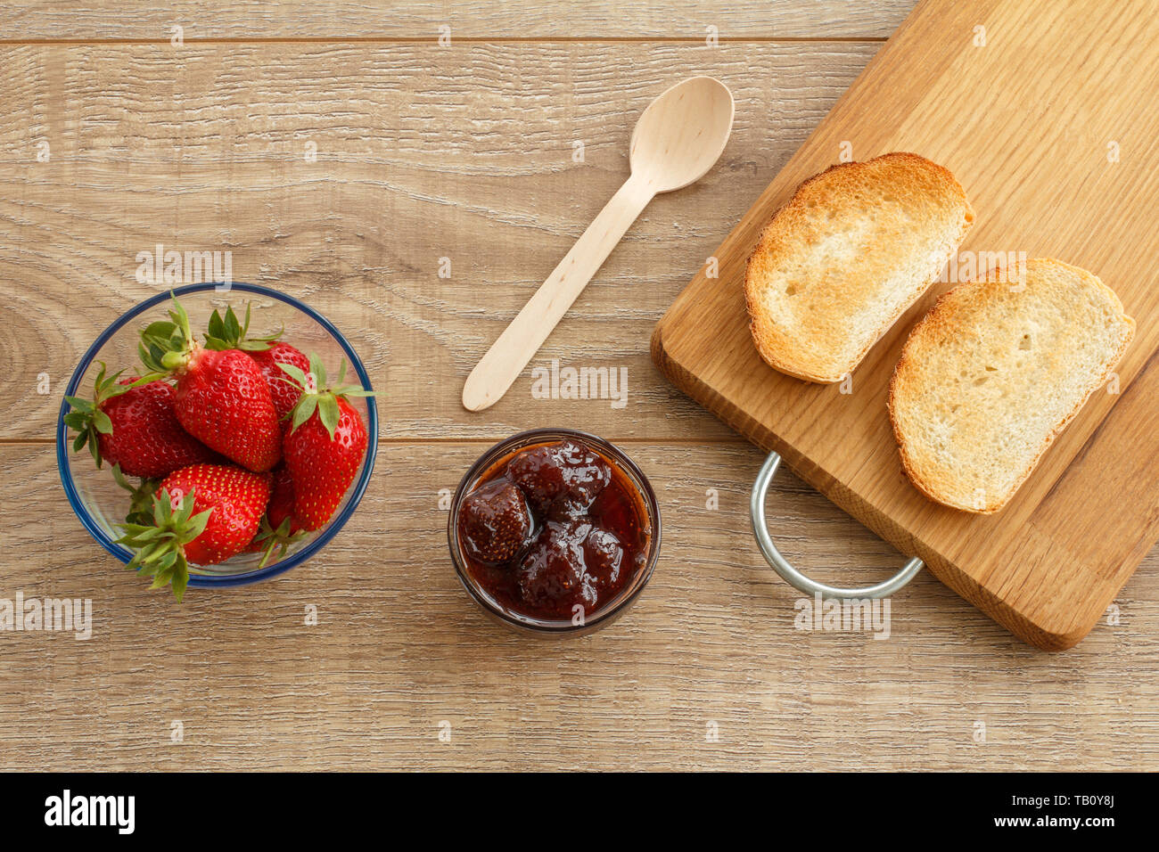 Traditional homemade strawberry jam in a glass bowl, fresh berries, toasts on cutting board with spoon on wooden desk. Top view. Stock Photo