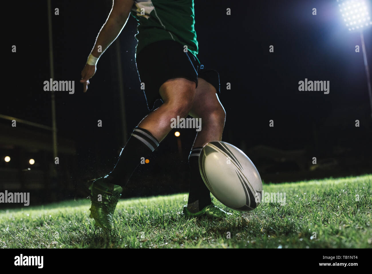 Rugby player drops the ball on the ground and then kicks it just as it bounces. Rugby player hitting a dropped goal under lights at sports arena. Stock Photo
