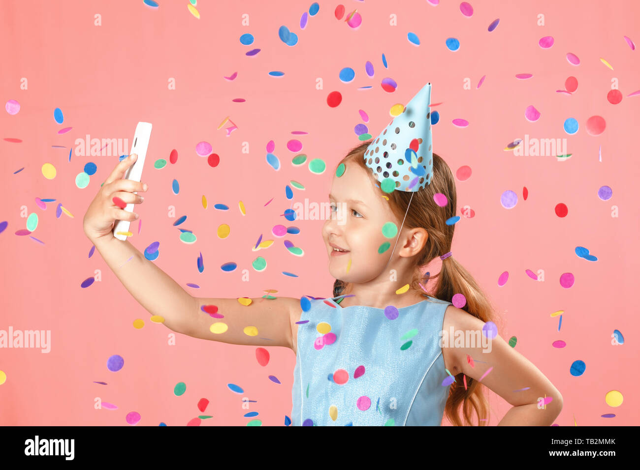 Cheerful little girl celebrates birthday. The child holds the phone, takes a selfie in the rain of confetti. Closeup portrait on pink background. Stock Photo