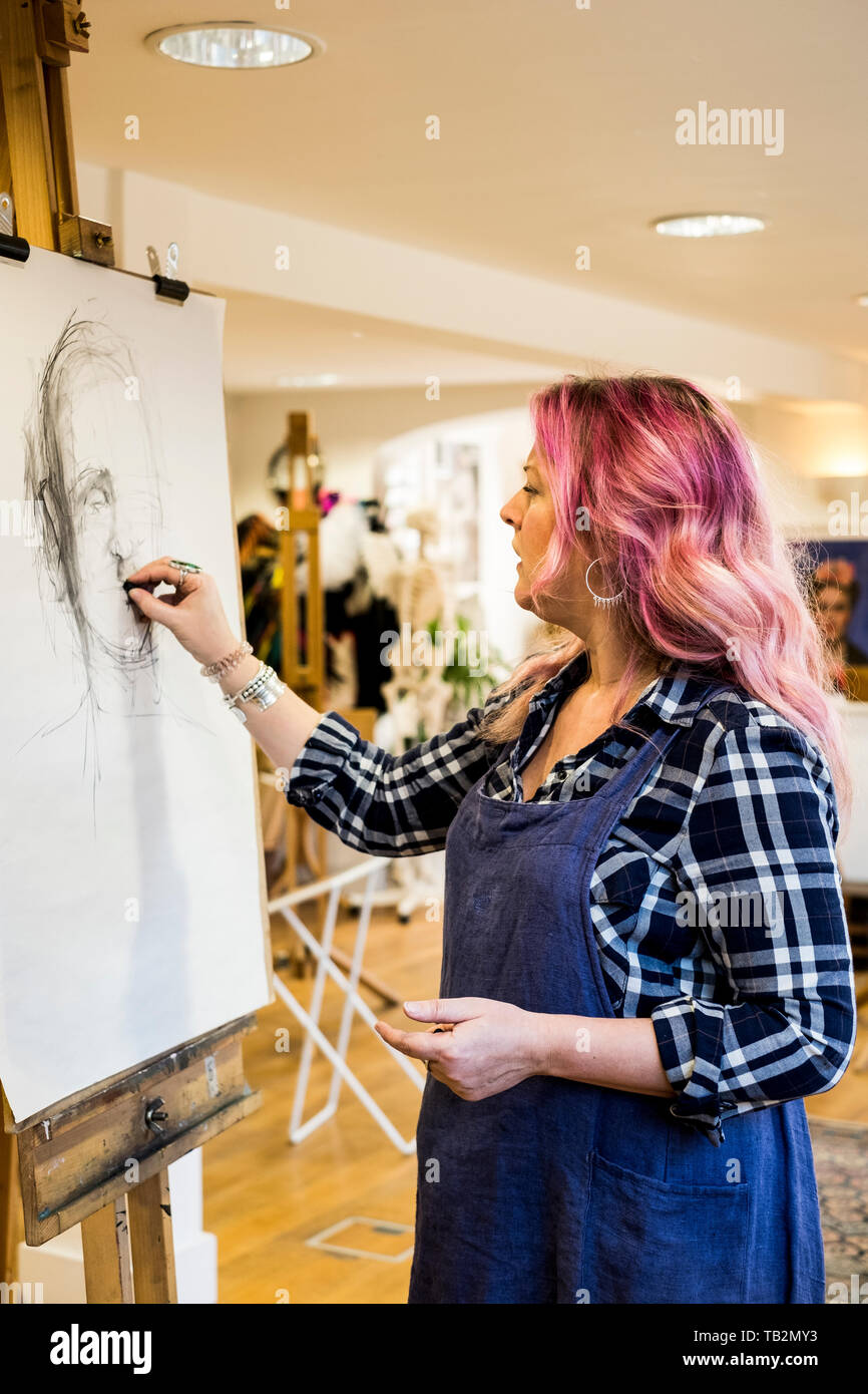 Woman with long blond wavy hair with pink streaks wearing apron standing at an easel, drawing a portrait. Stock Photo