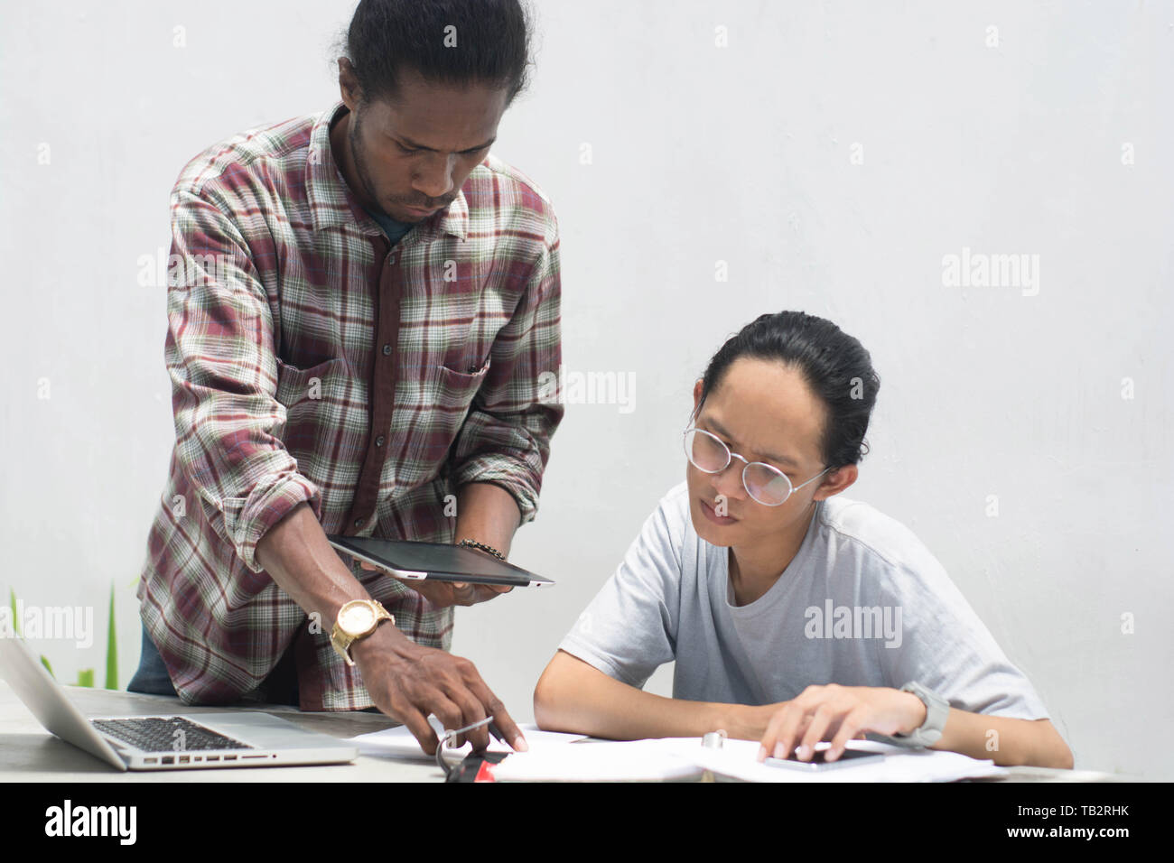 two friend with diffrent ethnic working together with laptop and tablet discussing something, two young man working and studying together Stock Photo