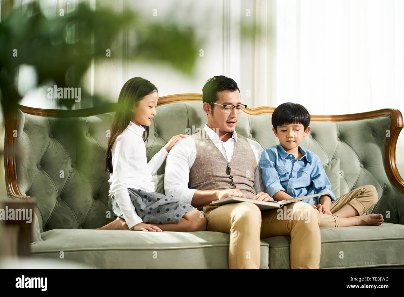 young asian father and two children sitting on couch reading book together in family living room at home Stock Photo