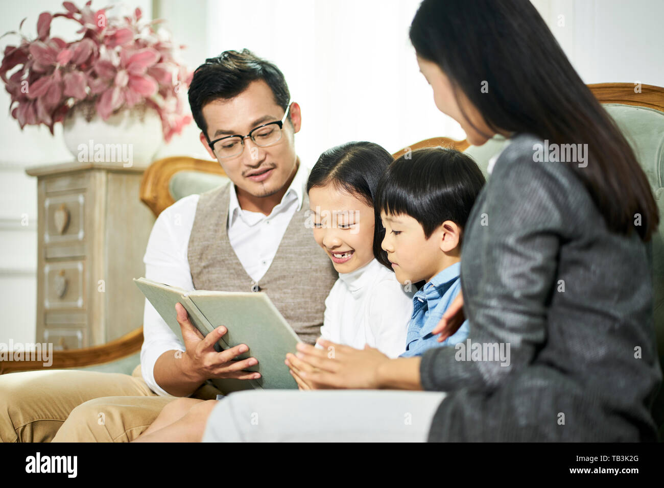 young asian parents and two children sitting on couch reading book together in family living room at home Stock Photo