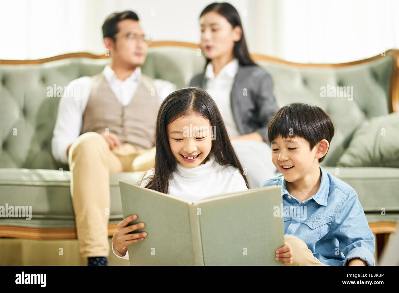 two asian kids brother and sister sitting on carpet reading book together in family living room with parents sitting on couch in the background. Stock Photo