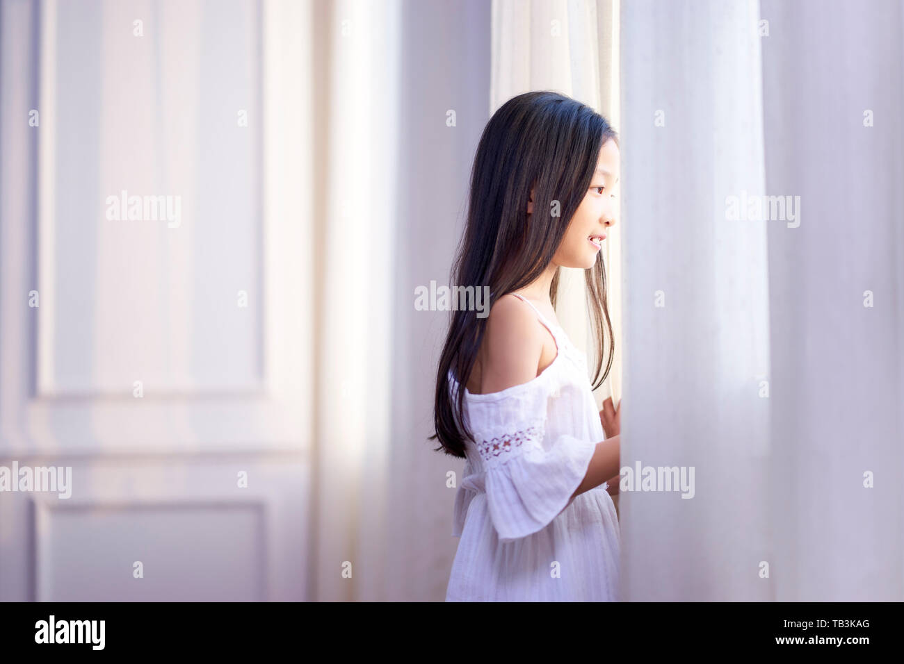 beautiful little asian girl with long black hair looking out of window in bedroom Stock Photo