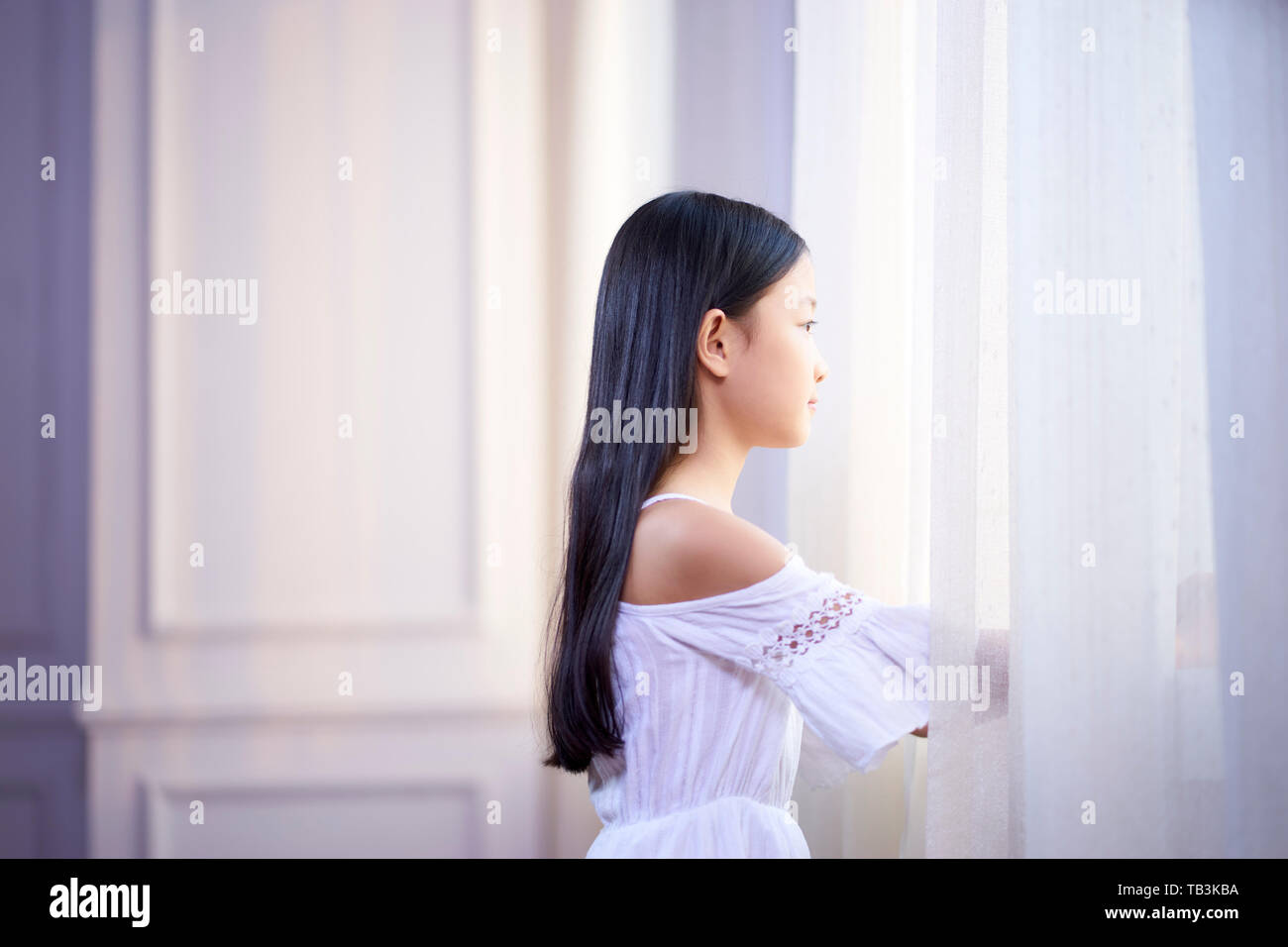 beautiful little asian girl with long black hair looking out of window in bedroom Stock Photo