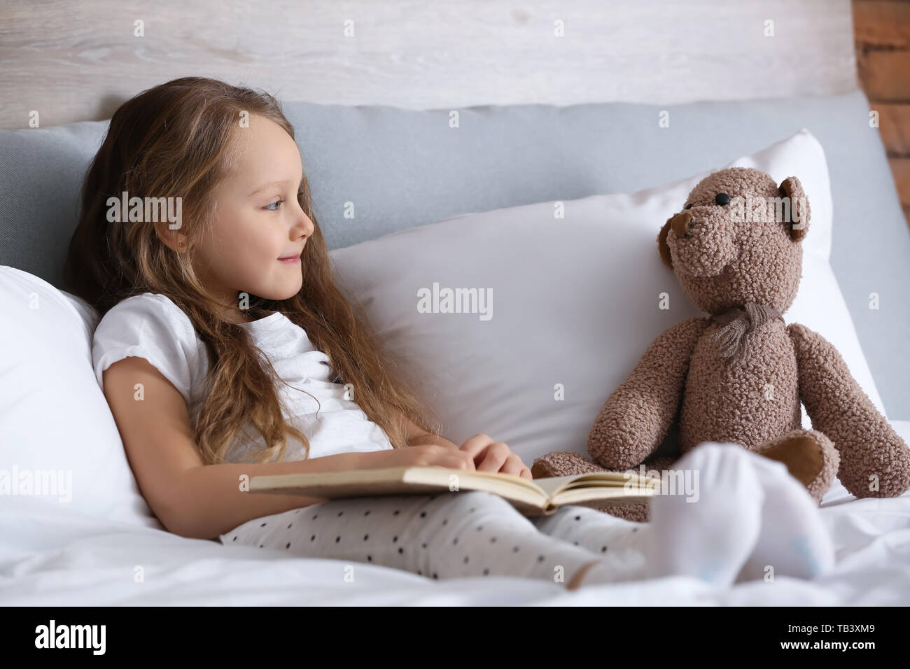 Cute little girl with toy bear reading book while sitting on bed Stock ...