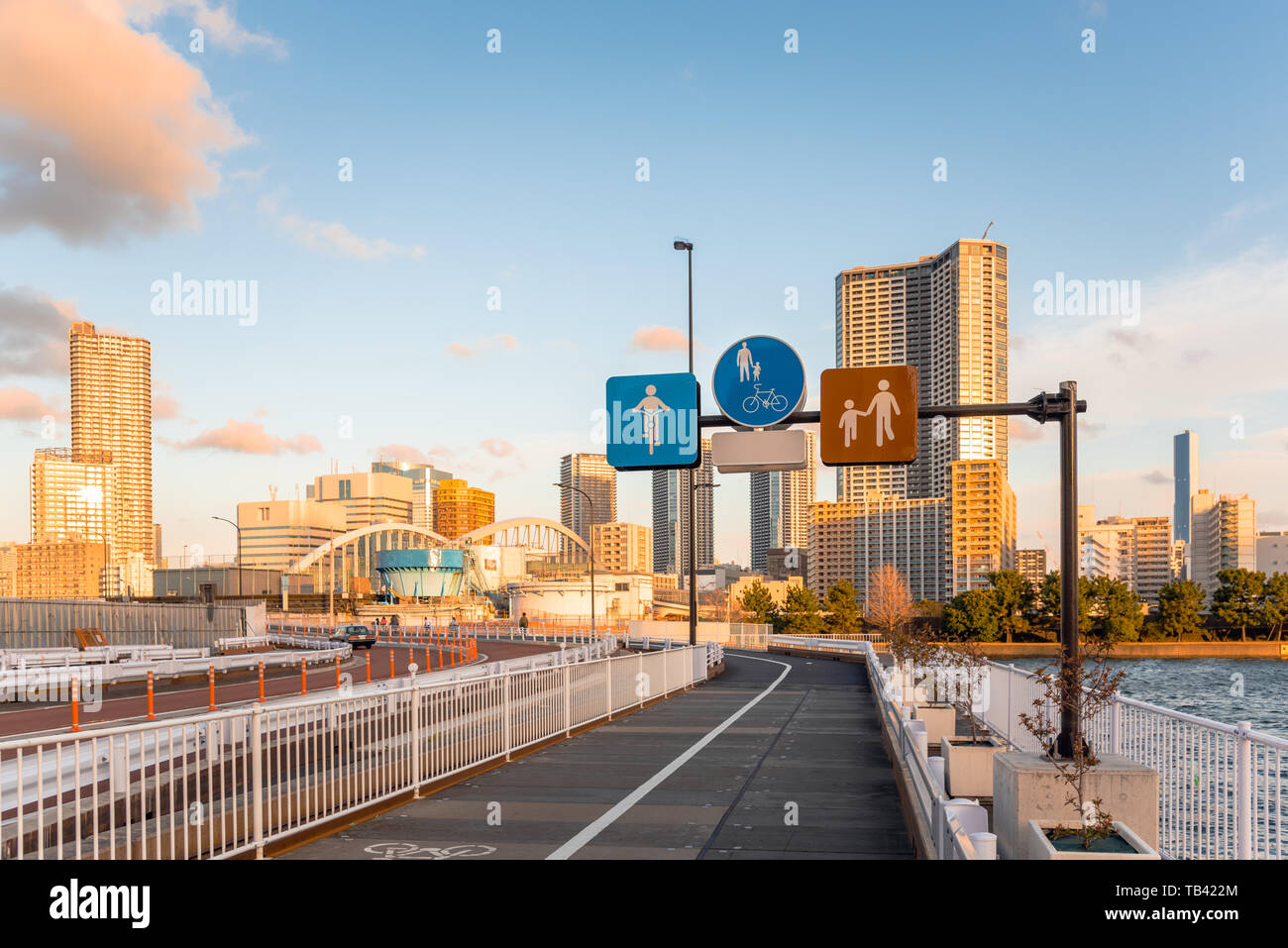 Road signs along a path for pedestrian and cyclists running alongside a street on Tokyo waterfront at sunset. Modern tall buildings arein background. Stock Photo