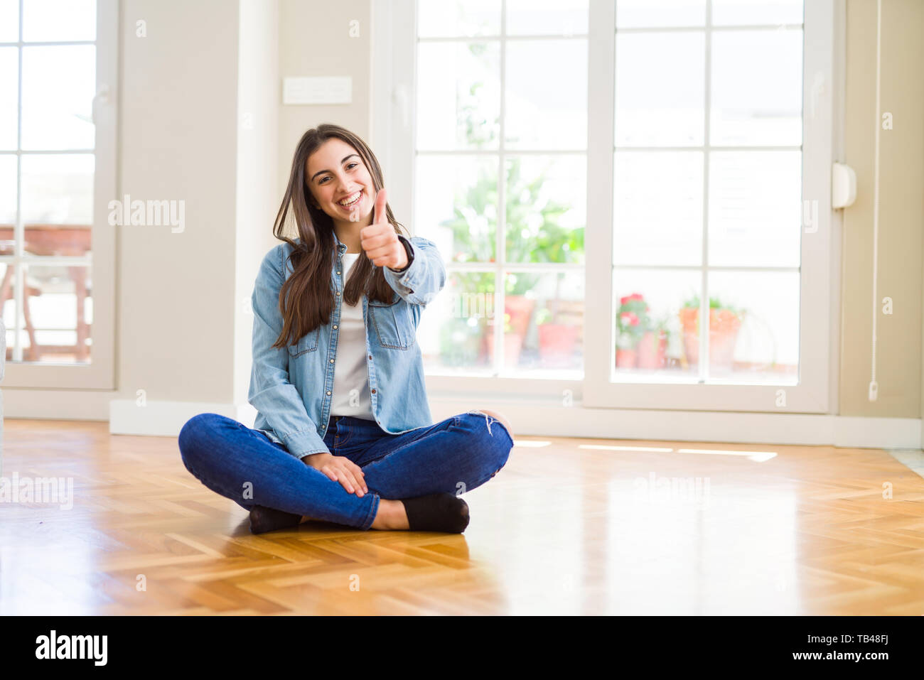 Beautiful young woman sitting on the floor at home doing happy thumbs ...