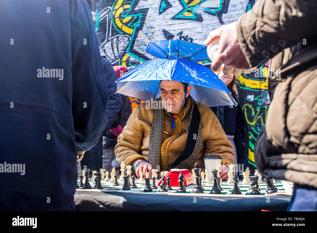Shoreditch, London, England, UK - April 2019: Old man street chess player playing chess wearing an umbrella as hat in Brick lane, East London Stock Photo
