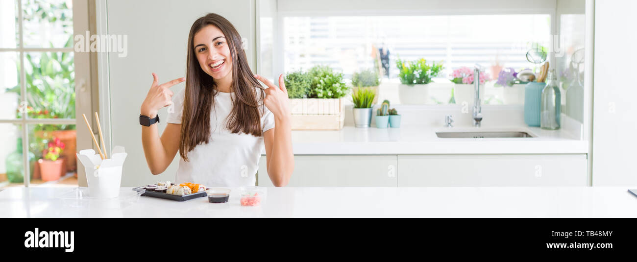 Wide angle picture of beautiful young woman eating asian sushi from delivery smiling confident showing and pointing with fingers teeth and mouth. Heal Stock Photo