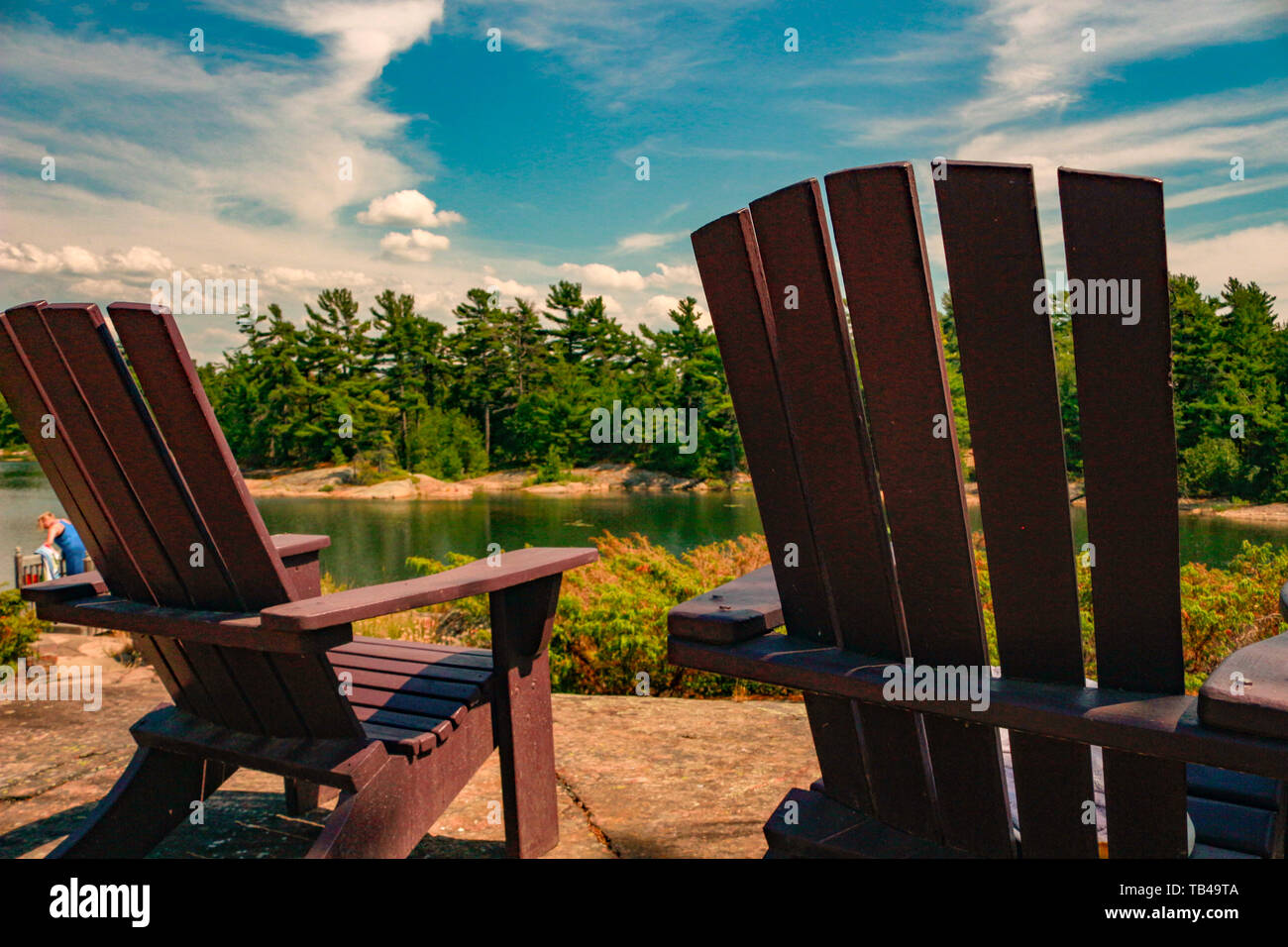 Two Muskoka chairs sitting on a wood dock facing a lake. Across the ...