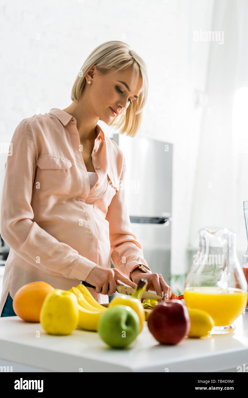 blonde pregnant young woman preparing food near fruits in kitchen Stock Photo