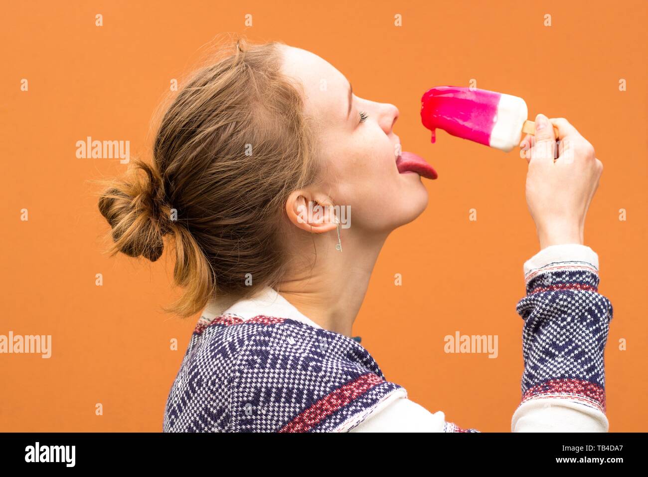 Beautiful and young girl eats a pink hue ice cream and enjoys. She catches a drop of melted ice cream with her tongue and smiles Stock Photo