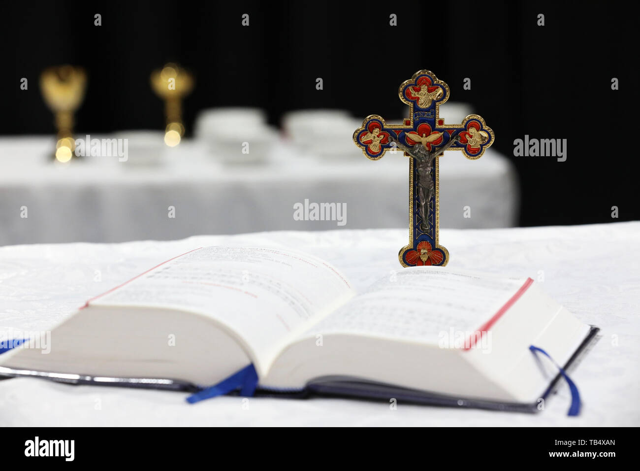 close up of open bible with holy crucifix behind it with jesus on the cross. Backround alter with blurred brass chalise and communion bowls. Stock Photo