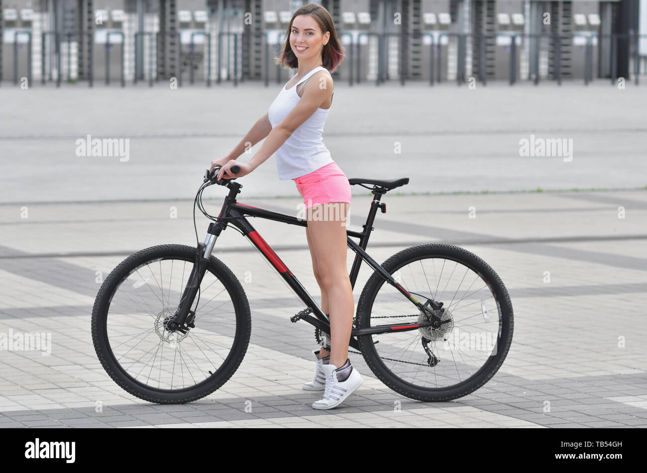 Young beautiful girl in sportswear stands next to a bicycle on the street in the city in summer Stock Photo